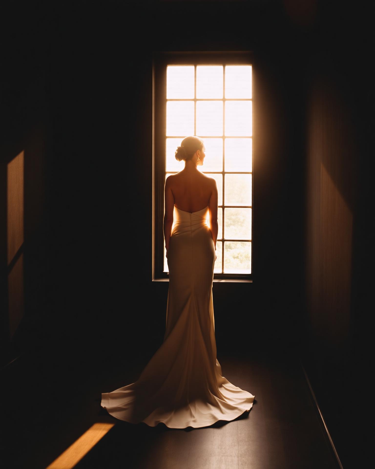 Bride in a structural ivory gown stands at a sunlit gallery window